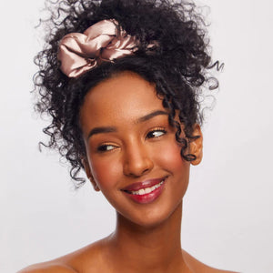 A package of KITSCH PILLOW SATIN SCRUNCHIES IN BLUSH sits behind four stacked scrunchies in beige, pink, cream, and champagne shades. The packaging displays a woman’s face and reads “Pillow Scrunchies in Satin” by KITSCH, showcasing this gentle, damage-free hair accessory ideal for soft styling.