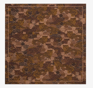 A person with long, wavy dark hair wears a light cream sweater and an intricately patterned brown fashion accessory from INOUI around their neck. The INOUI EDITIONS TETRIS SQUARE COTTON/SILK SCARF, made from soft cotton silk, features various shades of brown with detailed floral and geometric motifs. They stand against a plain white background.