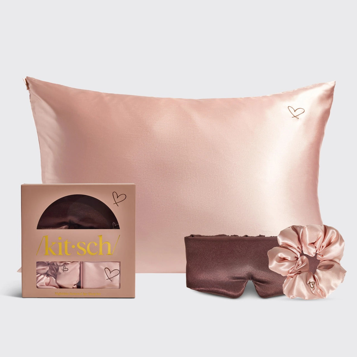 The KITSCH - NIGHTTIME RENEWAL 3PC SLEEP SET includes a pink satin pillowcase with a heart design, a chic boxed set with a brown satin eye mask, and a matching pink scrunchie, all elegantly showcased against a minimalist backdrop.