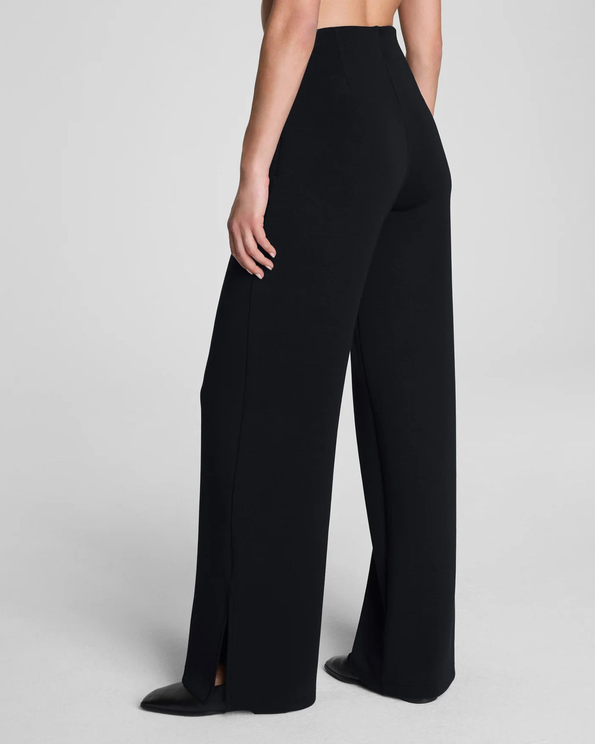 A person stands with one hand in their pocket, wearing SPANX AirEssentials Zero Waistband Wide Leg Pant made from light-as-air fabric and pointed black shoes against a plain light background.