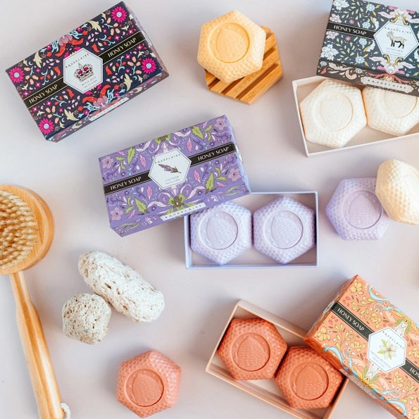 The BAUDELAIRE - LAVENDER HONEY SOAP 3.5 OZ, 2 BAR GIFT BOX features two hexagonal, triple-milled soaps with a beehive design, nestled in a decorative purple floral box and partially opened to reveal the lavender-colored bars inside.