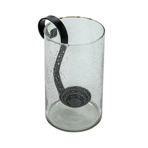 Three upright books are beside the CREATIVE COOP RECYCLED BUBBLE GLASS HURRICANE WITH METAL VOTIVE HOLDER, which contains a lit white candle and small pebbles. The lantern features a black metal handle and sits against a plain light background.