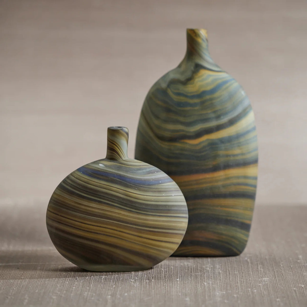 The ZODAX LUCCA MARBELIZED GLASS VASE - SHORT is a round, flat vase with a small off-center neck, featuring brown, tan, and blue marbled stripes, displayed on a white background.
