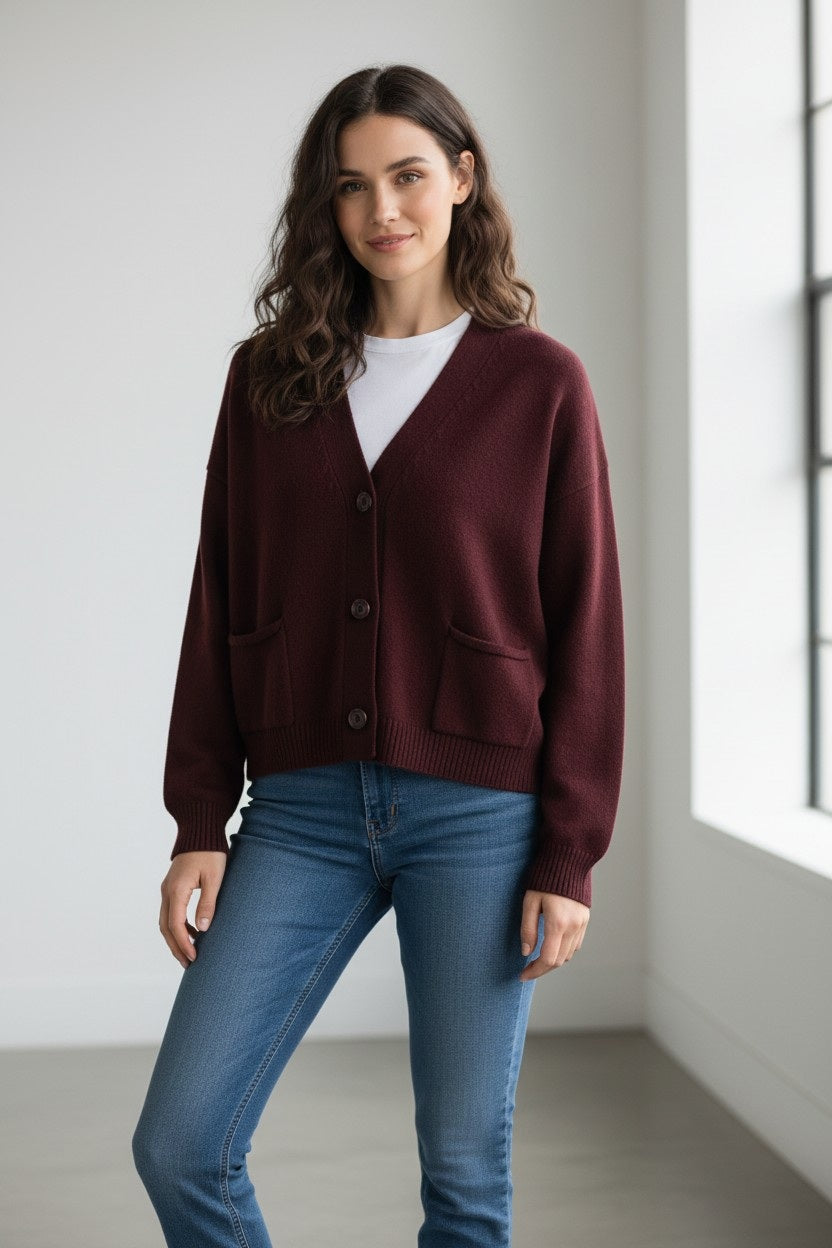 A woman with wavy brown hair stands indoors near a large window, smiling softly at the camera while wearing the FLOOR - SWEATER CARDIGAN WITH POCKETS by FLOOR, an oversized cropped wool cardigan made in Italy, layered over a white shirt and blue jeans.