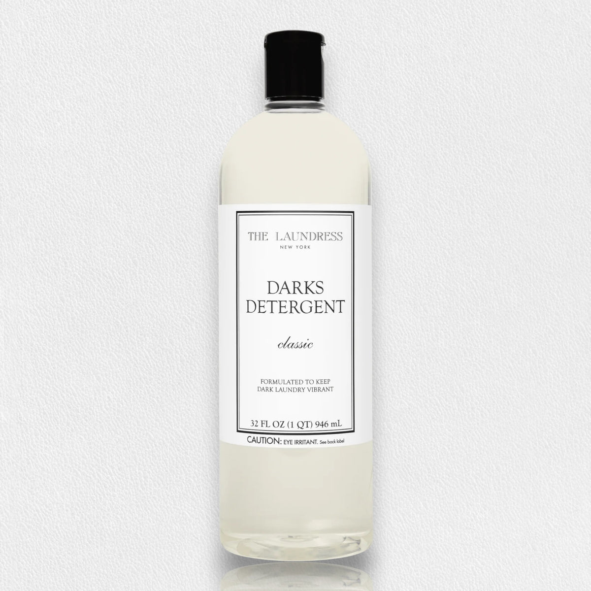 A clear bottle of THE LAUNDRESS - DARKS DETERGENT 32OZ with a black cap is displayed against a white background. The label reads &quot;Darks Detergent Classic, formulated with an enzyme blend to keep dark laundry vibrant, 32 fl oz (1 qt) 946 ml.&quot; A caution note is visible at the bottom.