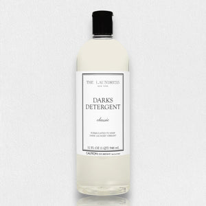 A clear bottle of THE LAUNDRESS - DARKS DETERGENT 32OZ with a black cap is displayed against a white background. The label reads "Darks Detergent Classic, formulated with an enzyme blend to keep dark laundry vibrant, 32 fl oz (1 qt) 946 ml." A caution note is visible at the bottom.