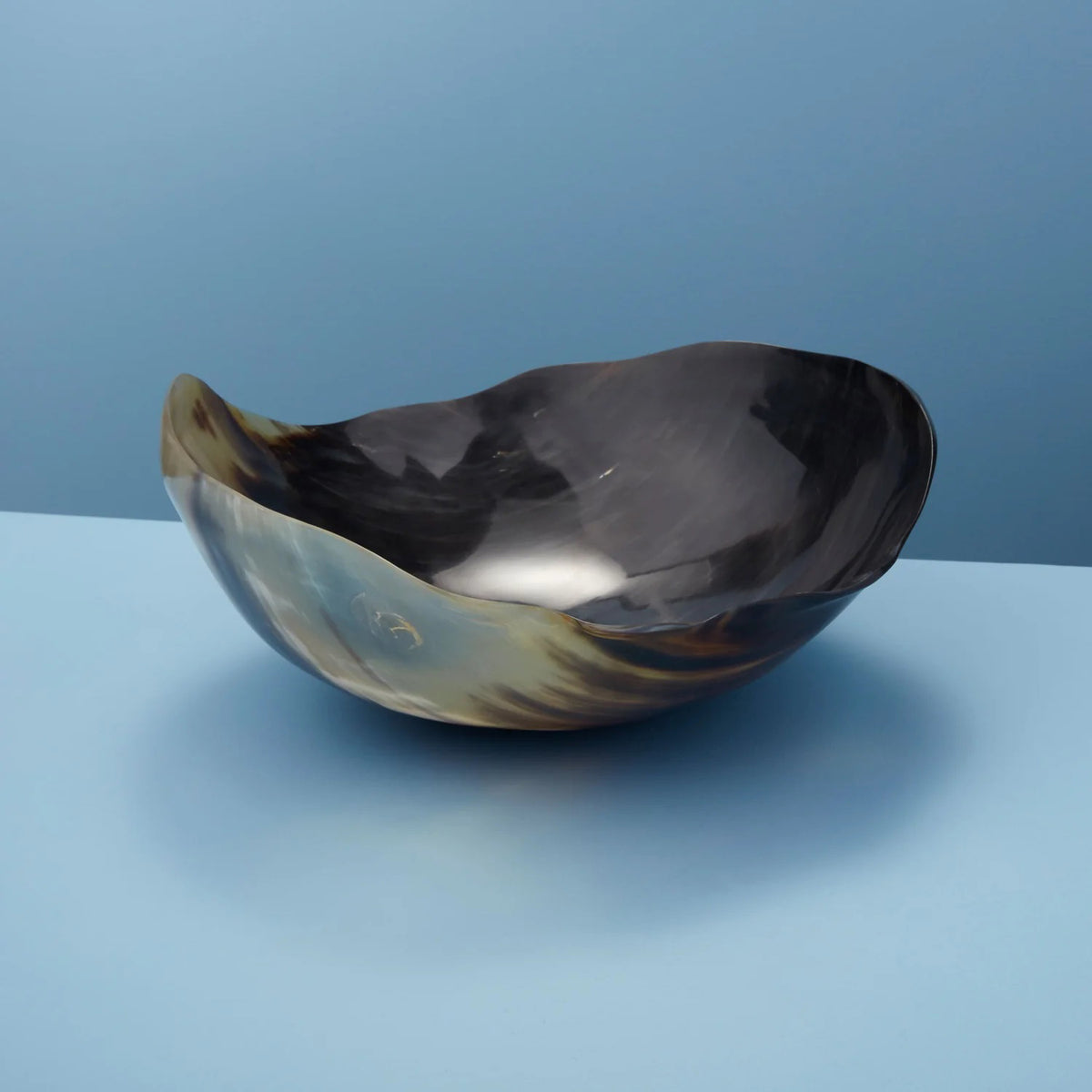 The HORN WAVY ROUND BOWL by BE HOME, INC, ethically crafted from water buffalo horn, showcases a polished, reflective surface. Its irregular, organic shape rests on a flat blue background. The natural blend of black and tan colors highlights the bowl&#39;s unique texture and sheen.