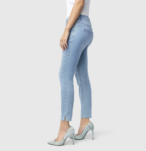 With long brown hair, a person stands against a plain backdrop wearing MAC's tan cropped jacket and MAC JEANS - DREAM SUMMER light blue slim jeans made from a comfortable modal blend. White heels complete the look as they gaze ahead with relaxed arms by their sides.