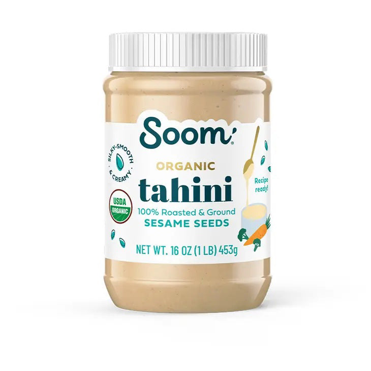 A jar of SOOM - ORGANIC TAHINI from SOOM FOODS, crafted entirely from silky-smooth, roasted and ground Ethiopian sesame seeds. The label proudly displays the USDA Organic certification and indicates a net weight of 16 ounces (453 grams). Additionally, it features an illustration of a spoon alongside ingredients such as carrots and cucumbers.