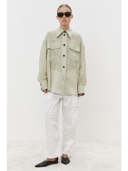 A person with brown hair in a low ponytail faces away, wearing an oversized MEIMEIJ - LINEN SHIRT. Against a plain white background, breathable linen pants are visible at the bottom.