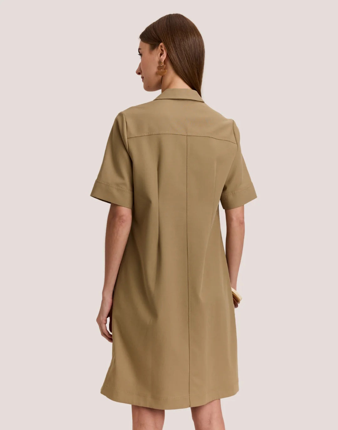 A woman wears the HINSON WU BRIDGET Short Sleeve Dress in Traveler’s Twill Khaki, featuring a collar and zip front. She pairs it with gold hoop earrings and bangles, standing against a plain light background.