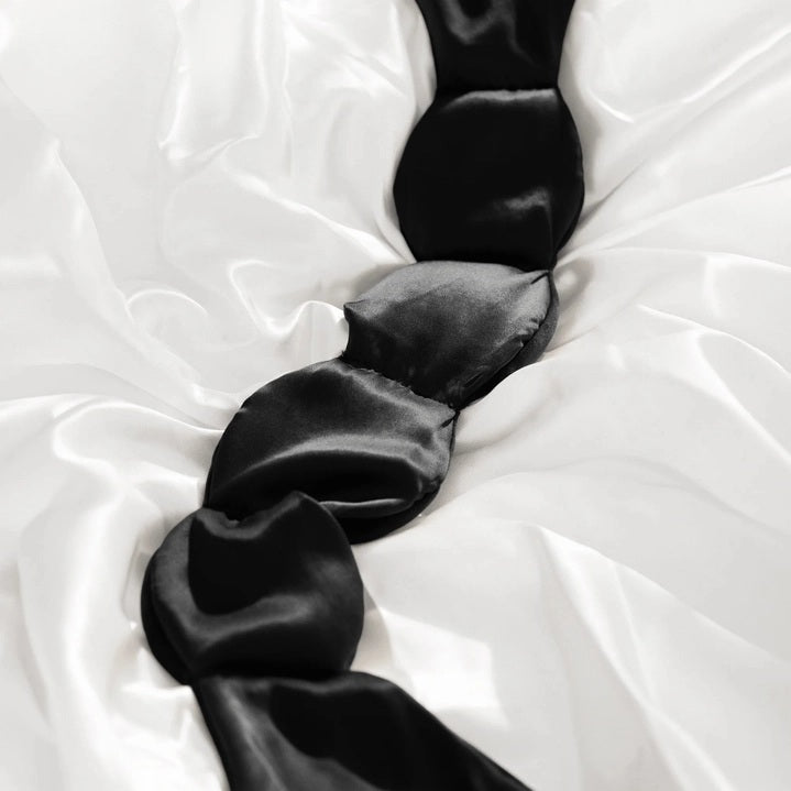 The NODPOD Silk Weighted Sleep Eye Mask in Noir by NODPOD is a dark blue, segmented eye mask featuring a strap-free design and unique four-pad shape. Text highlights its calming effect for stress relief and relaxation, all set against a light background.