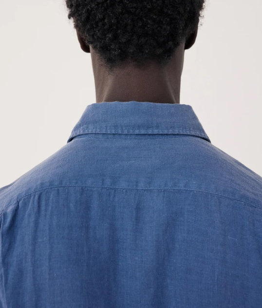 A man with short curly hair poses against a light background, wearing the HARTFORD - PAUL LINEN SHIRT IN DENIM made from European flax linen, featuring a chest pocket, paired with light pants. His right arm is bent behind his back.