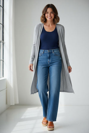 Someone stands against a neutral background wearing the MERSEA KATE WIDE LEG DENIM—light blue, high-waisted jeans made from recycled cotton—paired with a navy top, gray cardigan, and tan platform sandals.
