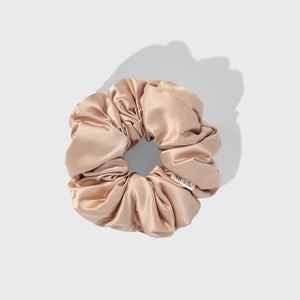 A package of KITSCH PILLOW SATIN SCRUNCHIES IN BLUSH sits behind four stacked scrunchies in beige, pink, cream, and champagne shades. The packaging displays a woman’s face and reads “Pillow Scrunchies in Satin” by KITSCH, showcasing this gentle, damage-free hair accessory ideal for soft styling.
