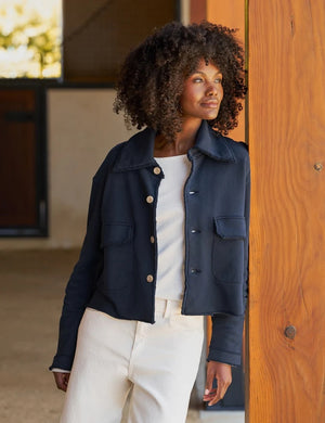 A woman with long brown hair wearing the FRANK & EILEEN Rowan Cropped Sweatshirt Utility Jacket in Triple Terry Classic Navy, paired with white pants, stands sideways and smiles outside near a white building with windows.
