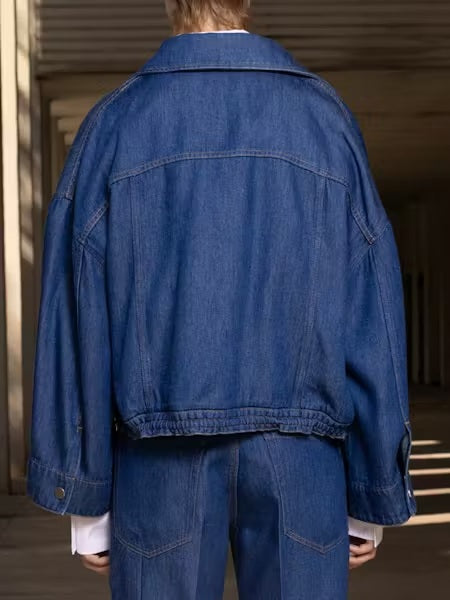 A person with straight brown hair wears the NUDE - DENIM JACKET by NUDE, layered over a white button-up shirt and blue jeans, standing against a plain light background.