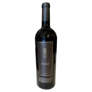 A bottle of LONG SHADOWS SAGGI SANGIOVESE 2021 with a black label featuring an owl illustration and the text “Saggi 2021,” standing on a white surface in front of wine shelves.