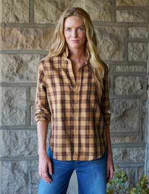 A woman stands in a sunlit room, smiling with curly hair, wearing the FRANK & EILEEN EILEEN Relaxed Button Up Shirt in Italian Flannel Gold Brown Plaid, paired with blue skinny jeans and brown ankle boots.