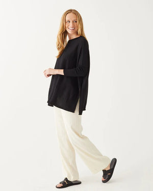 A smiling woman with long blonde hair wears the MERSEA Catalina Crewneck Sweater in black, featuring signature split sides, paired with light pants and standing against a plain white background.
