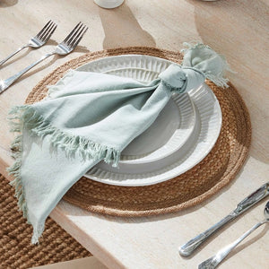 A stack of NAPA HOME AND GARDEN - AURA DINNER NAPKIN IN LIGHT GREEN, featuring fringed edges, is folded and placed on a white surface.