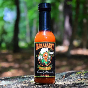 A 5 fl oz (150 ml) bottle of CACKALACKY - JACK PEPPER SAUCE GOLD by CACKALACKY, featuring a black label with a cartoon pepper in a chef’s hat, plus white, orange, and green text. This is an all-natural hot sauce.
