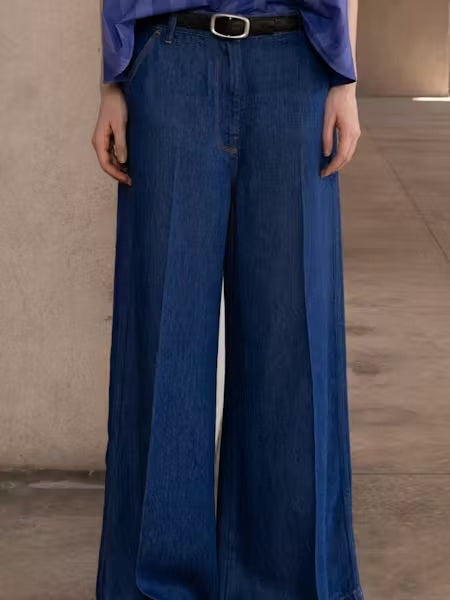 A person stands indoors, shown from the waist down, wearing NUDE's TILDA WIDE LEG DENIM TROUSER with an ultra wide-leg silhouette covering their shoes, set against beige walls and a tiled floor.