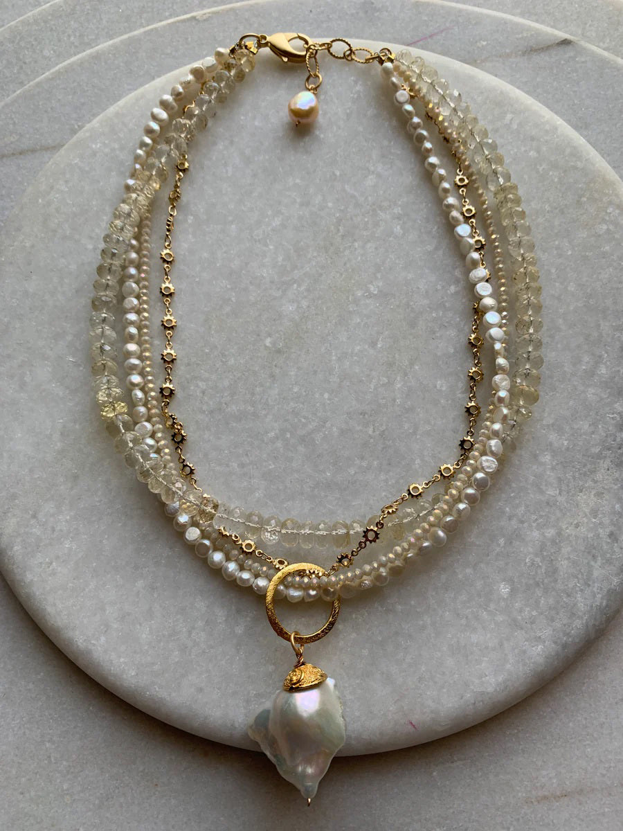 The BITTERSWEET DESIGNS - IVORY CRYSTAL FLANEUR necklace features layered gold chains, white pearls, translucent beads, and a large irregular pearl pendant—an exquisite handcrafted piece from BITTERSWEET DESIGNS displayed on a light stone surface.