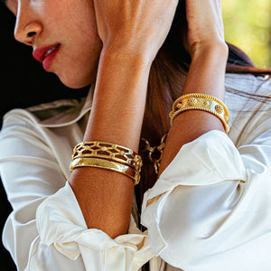A woman in a white blouse crosses her arms, showcasing several gold bracelets on her wrists, including the bold CAPUCINE DE WULF BERRY CLASSIC CUFF. The softly blurred background features green and yellow tones.
