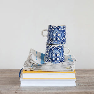The CREATIVE COOP CREAM AND BLUE HAND-PAINTED STONEWARE MUG features a blue floral and leaf design on white, a handle, tapered shape, and glossy finish—bringing artisanal charm to your daily coffee.