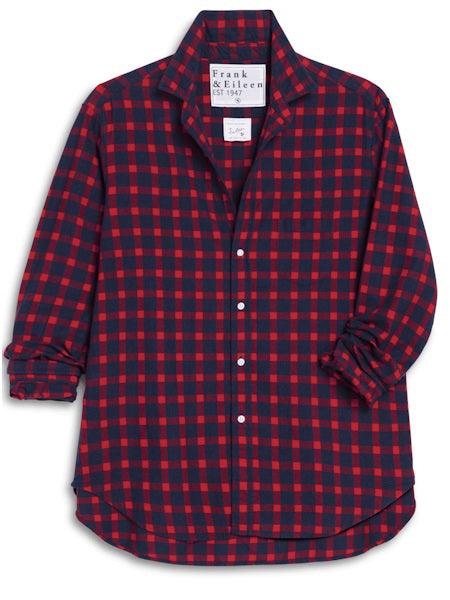 A blonde woman with a ponytail stands indoors, smiling and wearing the FRANK &amp; EILEEN EILEEN Relaxed Button Up Shirt in Italian Flannel Red Blue Plaid. Sunlight pours through glass doors, highlighting a nearby potted plant.