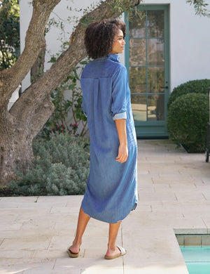 A person stands outdoors on a patio, wearing a FRANK & EILEEN Rory Maxi Shirtdress in Vintage Stoneash Denim and white sandals. They have curly hair, are looking to the side with one hand in a pocket, surrounded by plants and a glass door.