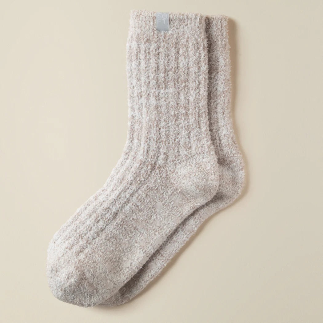 A pair of feet wear SOFTIES' Marshmalow Crew Socks in Heather Coco, raised against a beige background. These plush, cozy socks cover the ankles and add comfort with their soft, textured design.