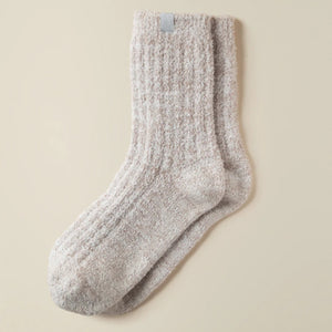 A pair of feet wear SOFTIES' Marshmalow Crew Socks in Heather Coco, raised against a beige background. These plush, cozy socks cover the ankles and add comfort with their soft, textured design.