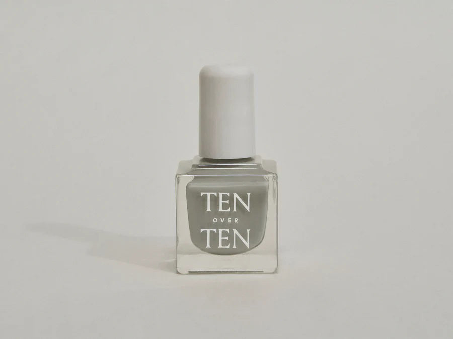 A glass bottle of TENOVERTEN - NAIL POLISH from the brand TEN OVER TEN, featuring a white cap and a dusty pink nail color. The front label displays "TEN OVER TEN" in white uppercase letters. This vegan, eight-free formula ensures both beauty and peace of mind. The background is plain and light-colored.