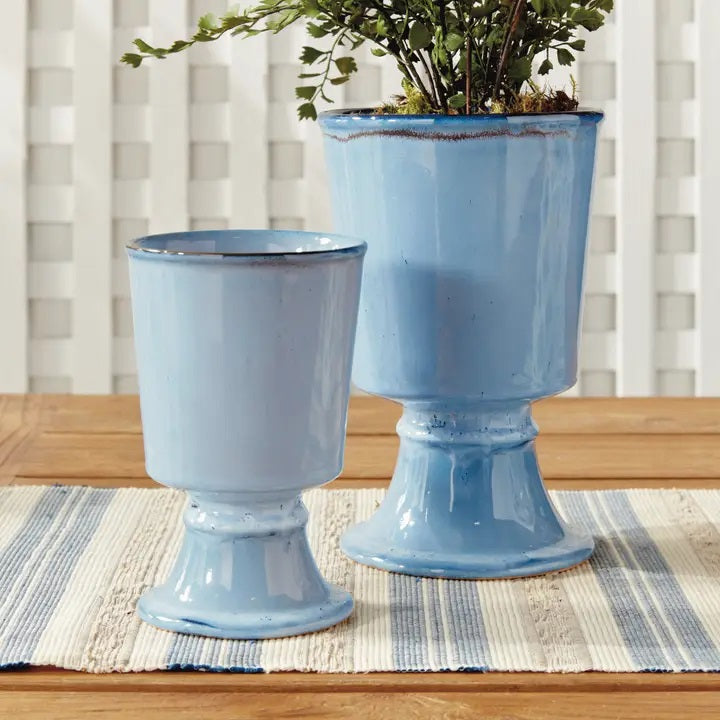 The NAPA HOME AND GARDEN Blue Kelcy Footed Cachepot, Small features a light blue ceramic finish with a glossy reactive glaze, subtle darker speckles near the rim and base, plus a wide rim and sturdy flared foot for a classic look.