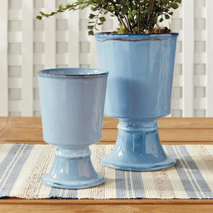 The NAPA HOME AND GARDEN Blue Kelcy Footed Cachepot, Small features a light blue ceramic finish with a glossy reactive glaze, subtle darker speckles near the rim and base, plus a wide rim and sturdy flared foot for a classic look.