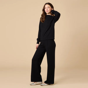 A woman wears a SOFTIES - DREAMTECH BOAT NECK WITH PINTUCK PANT SET in olive green, featuring a long-sleeve boat neck top and matching wide-leg pintuck pants.