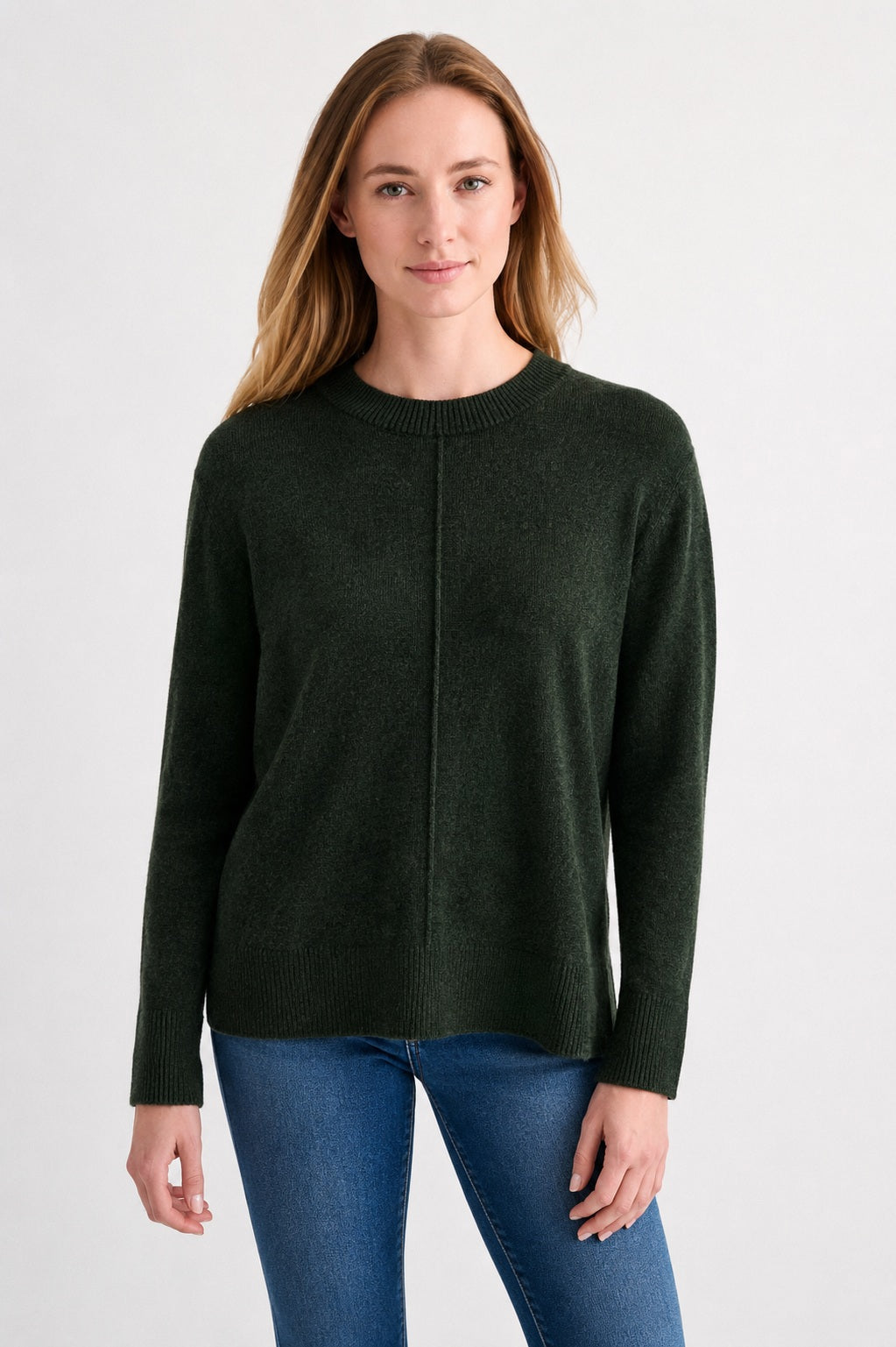 A woman with long, light brown hair wears the KINROSS CASHMERE - HI LOW SEAMED CREW NECK CASHMERE SWEATER IN HIGHLAND and blue jeans. She stands facing forward against a plain white background, smiling slightly.
