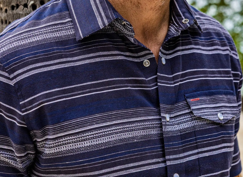 A man smiles with one hand in his pocket, wearing the CRIQUET Hemp Stripe Pearl Snap Shirt in Navy—short-sleeve, button-up with white stripes—paired with beige pants.
