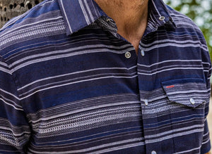 A man smiles with one hand in his pocket, wearing the CRIQUET Hemp Stripe Pearl Snap Shirt in Navy—short-sleeve, button-up with white stripes—paired with beige pants.