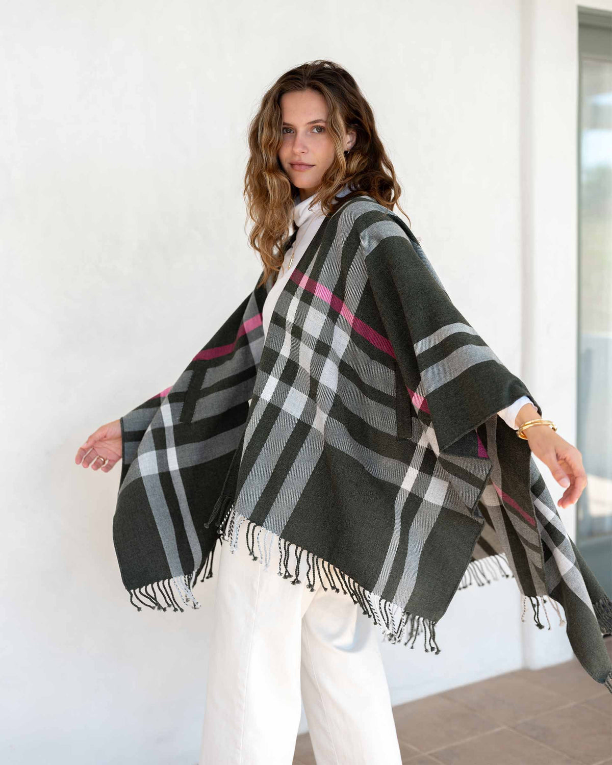 A woman with wavy brown hair wears the MERSEA Plaid Pocket Wrap—a black, white, and red plaid poncho with fringed edges—over a white outfit indoors. This MERSEA travel wrap is perfect for cozy style on the go.