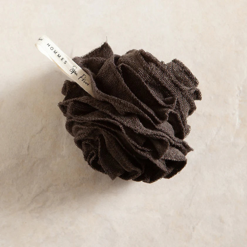 A black bamboo pouf, an eco-friendly bath accessory, nestled inside grey and gold packaging. The label reads &quot;EUROPEAN SOAPS, LLC&quot; with a sleek design that includes geometric patterns and the words &quot;Hommes&quot; and &quot;Spa Privé&quot;.