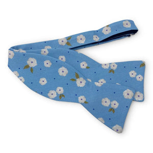 The COLLARED GREENS - COROLLA CAROLINA COTTON BOW TIE IN BLUE by BIRD DOG BAY features a light blue background with small white flowers, green leaves, and scattered blue dots in a handmade design.