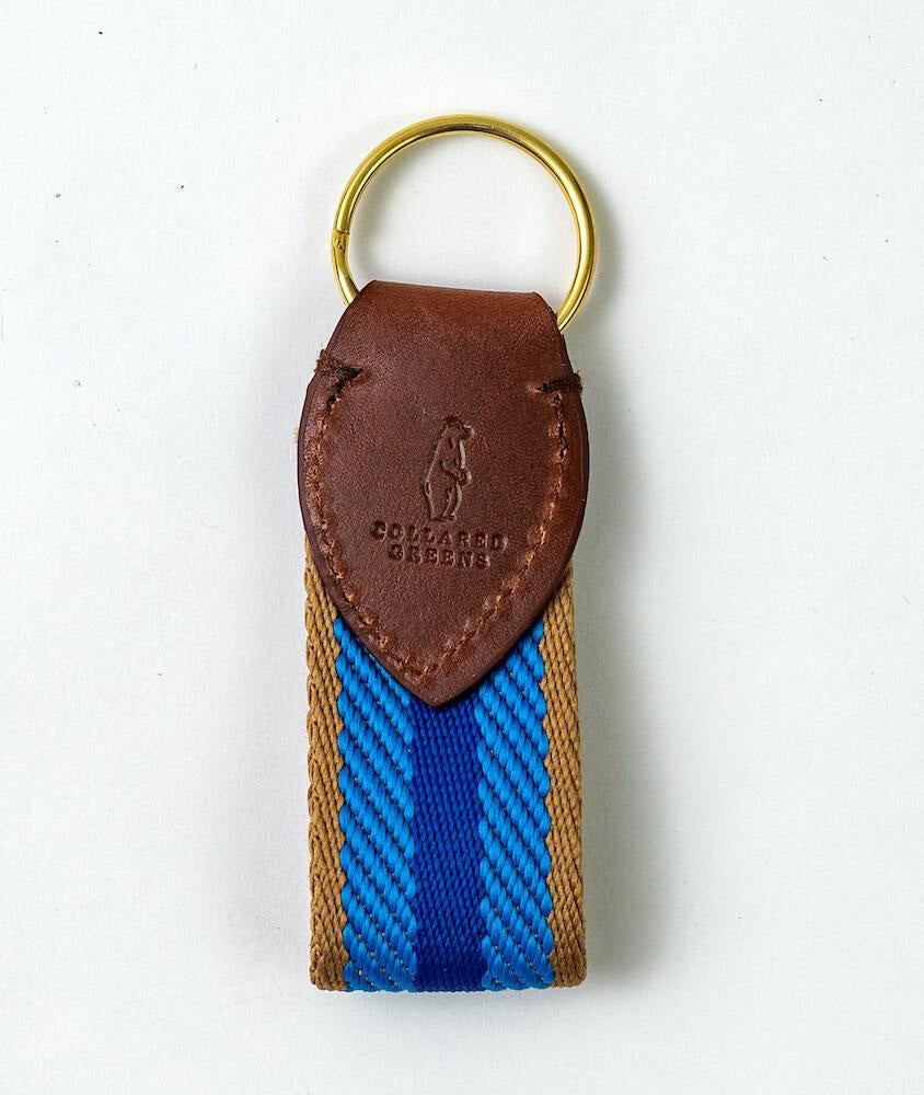 The BIRD DOG BAY Collared Greens Dockside Keychain in navy, light blue, and tan features a gold metal ring, brown leather top embossed with the logo, and a striped fabric strap—Made in America and shown on a white background.
