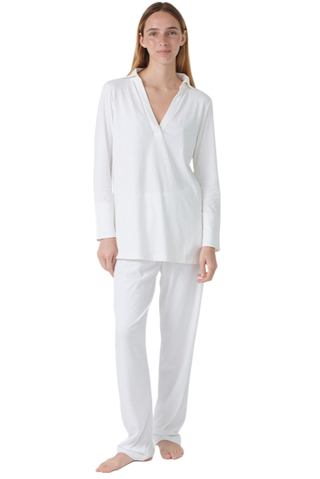 A person stands barefoot against a white backdrop, subtly smiling in the FASHIONVIEWS PJAMAS - BUTTERKNIT SPORTY PAJAMA, a white long-sleeved set crafted from soft pima cotton.