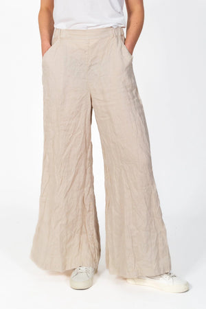 A pair of CP SHADES - WENDY PALAZZO PANT in beige is hanging on a wooden hanger against a light gray background. These cotton pants from CP SHADES feature an elastic waistband and handy side pockets, designed with a high-rise and wide-leg silhouette.