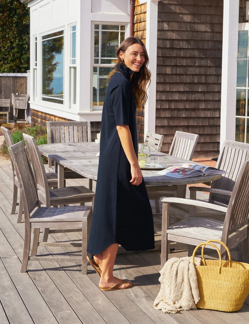A woman in the FRANK &amp; EILEEN EMMA Popover Henley Maxi Dress in British Royal Navy and sandals walks on a wooden deck beside an outdoor dining table, with a yellow straw bag and blanket nearby, and a house with large windows in the background.