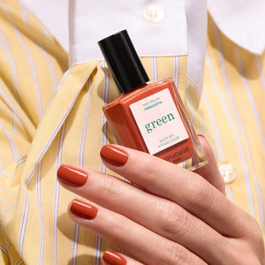 A hand with a glossy coat of MANUCURIST - GREEN™ NAIL POLISH IN TERRACOTTA by MANUCURIST INC touches an ear adorned with a chunky, abstract silver earring against brown skin.