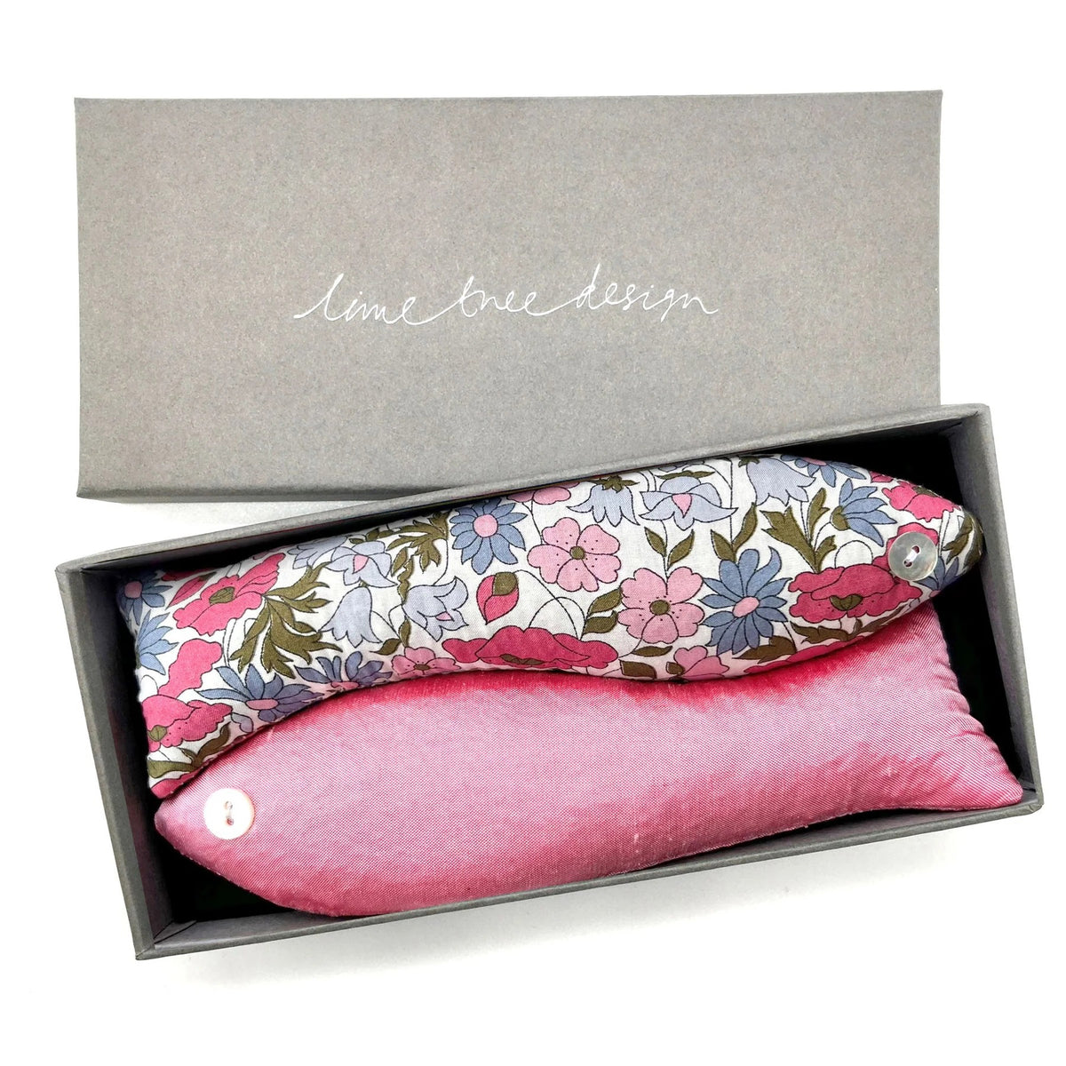 The LAVENDER FISH AND CINDERELLA SACHET set from LIME TREE COLLECTION LTD features a rectangular grey gift box that includes two delightful handmade fabric fish. One fish showcases a floral pattern with pink, white, and blue flowers, while the other is solid pink. The lid of the box, placed to the side, displays the text &quot;LIME TREE DESIGN.&quot; These charming lavender fish make for an ideal gift.